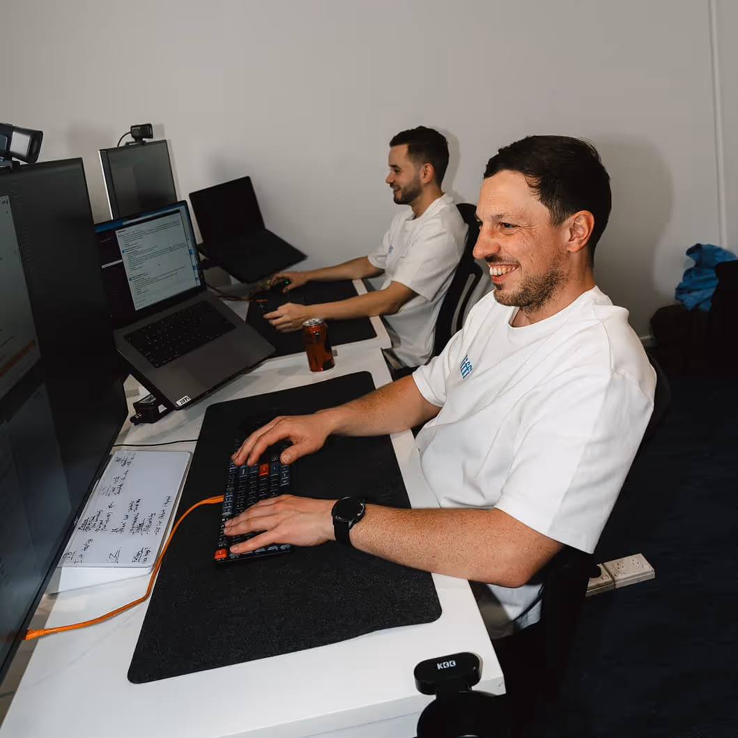 Two men working at desks with multiple computer screens, one smiling and typing on a keyboard.