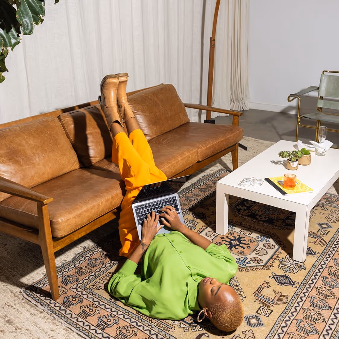 Person in a bright green shirt and orange pants lying on a patterned rug typing on a laptop near a brown leather couch and white coffee table.