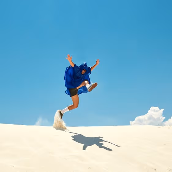 Person in blue poncho jumping energetically on a sand dune against a clear blue sky with a few clouds.