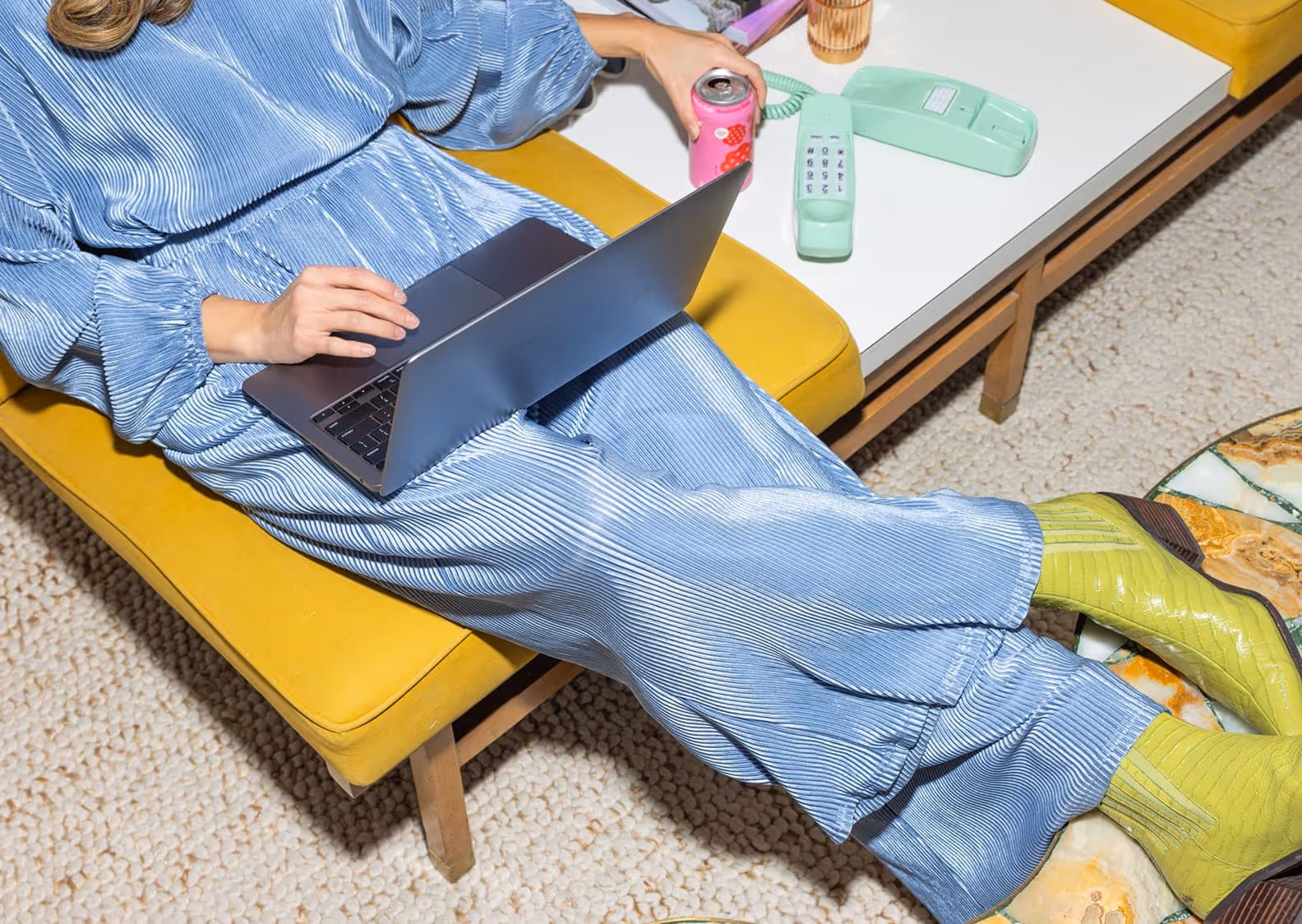Person in a blue pleated outfit sitting on a yellow bench using a laptop and holding a pink soda can near a white table with a mint green vintage telephone.