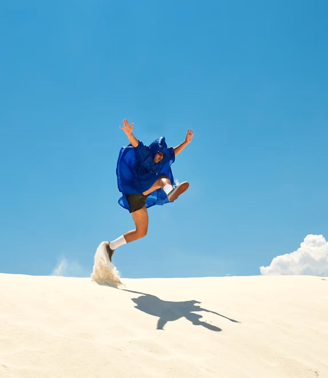 Person in blue poncho jumping energetically on a sand dune against a clear blue sky with a few clouds.