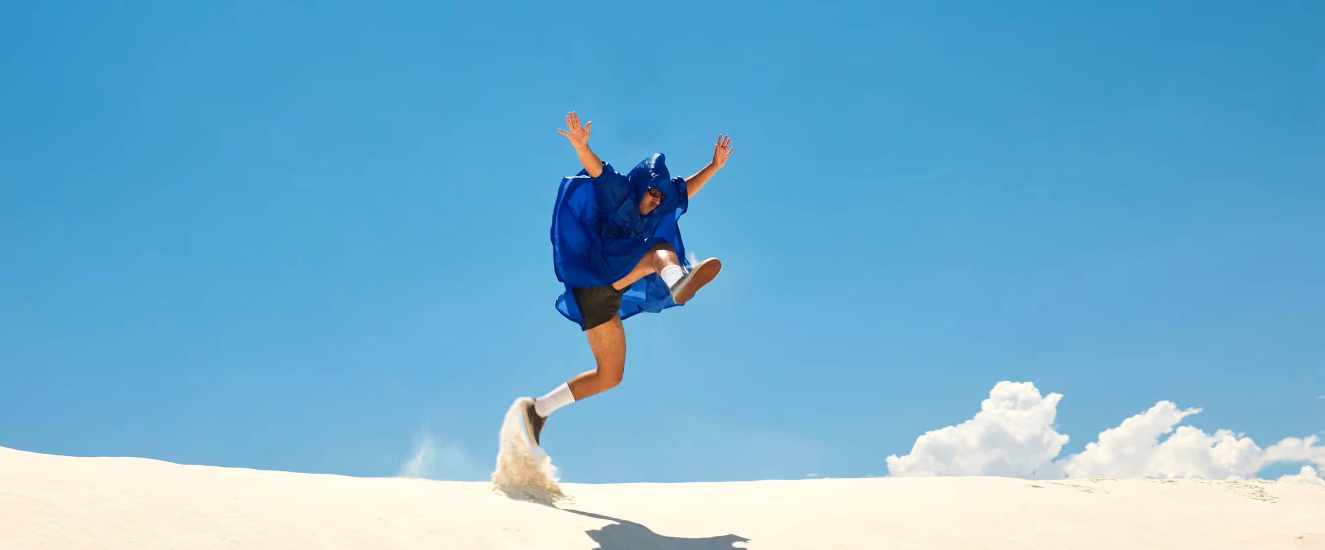 Person in blue poncho jumping energetically on a sand dune against a clear blue sky with a few clouds.