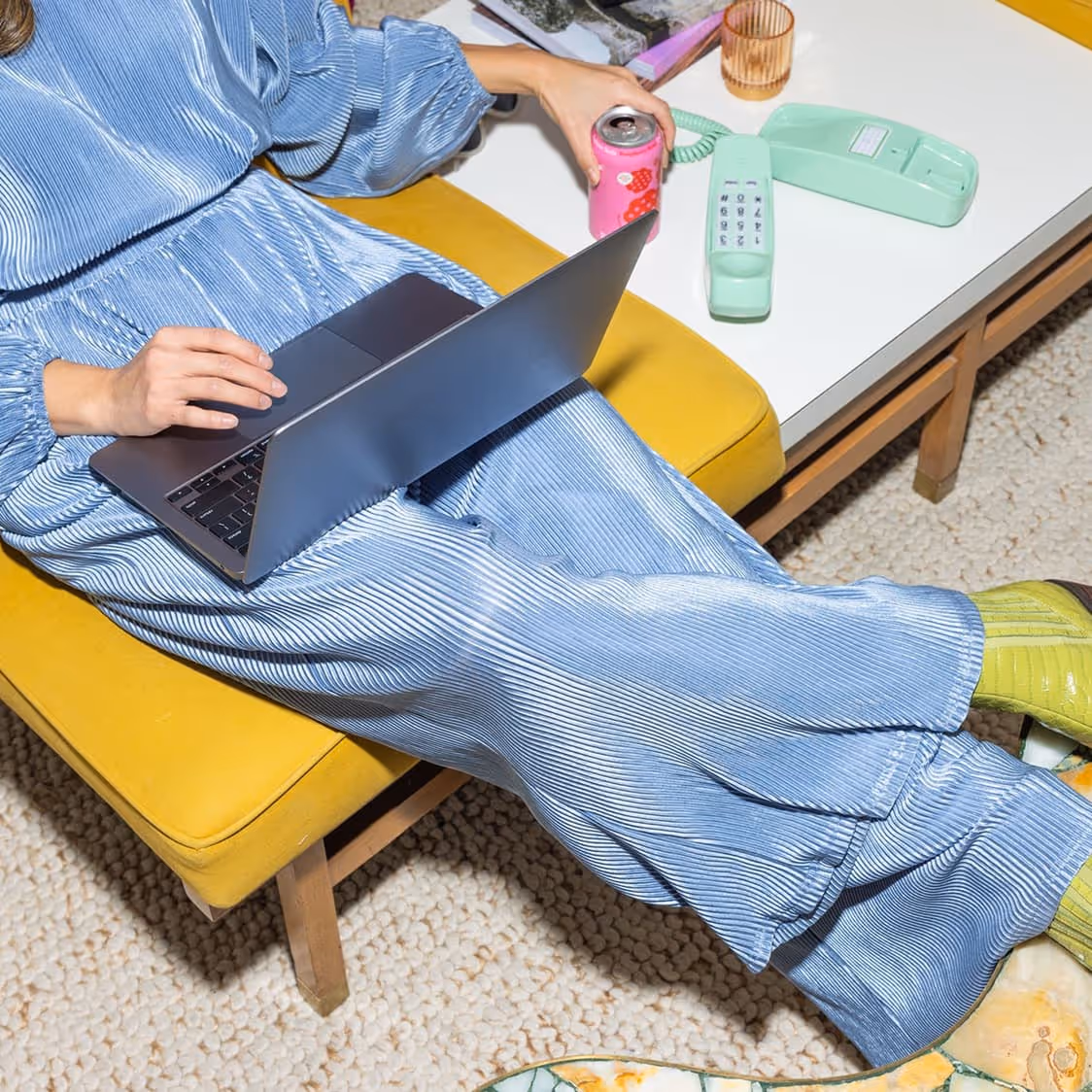 Person in a blue pleated outfit sitting on a yellow bench using a laptop and holding a pink soda can near a white table with a mint green vintage telephone.