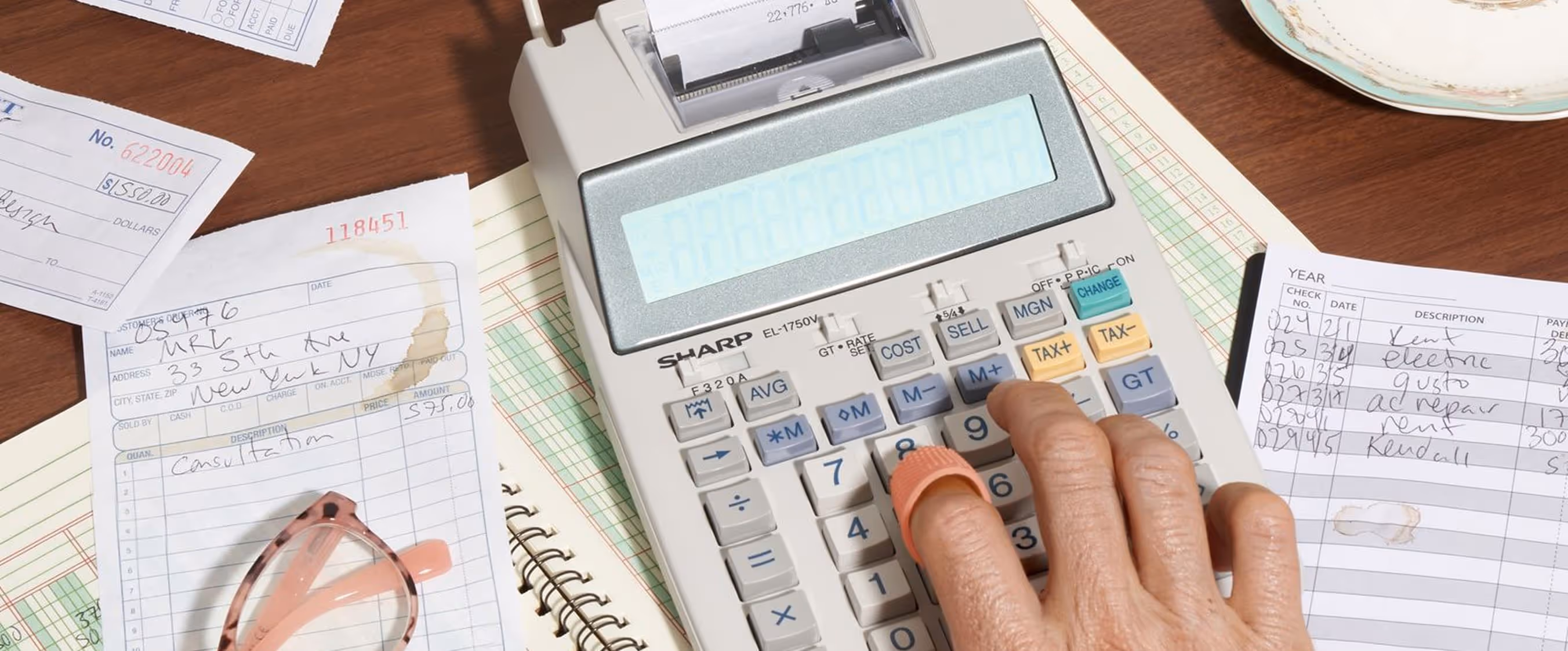A person’s hand uses a calculator on a desk with a ledger, receipts, glasses, a pen, and a cup of tea beside a computer keyboard.