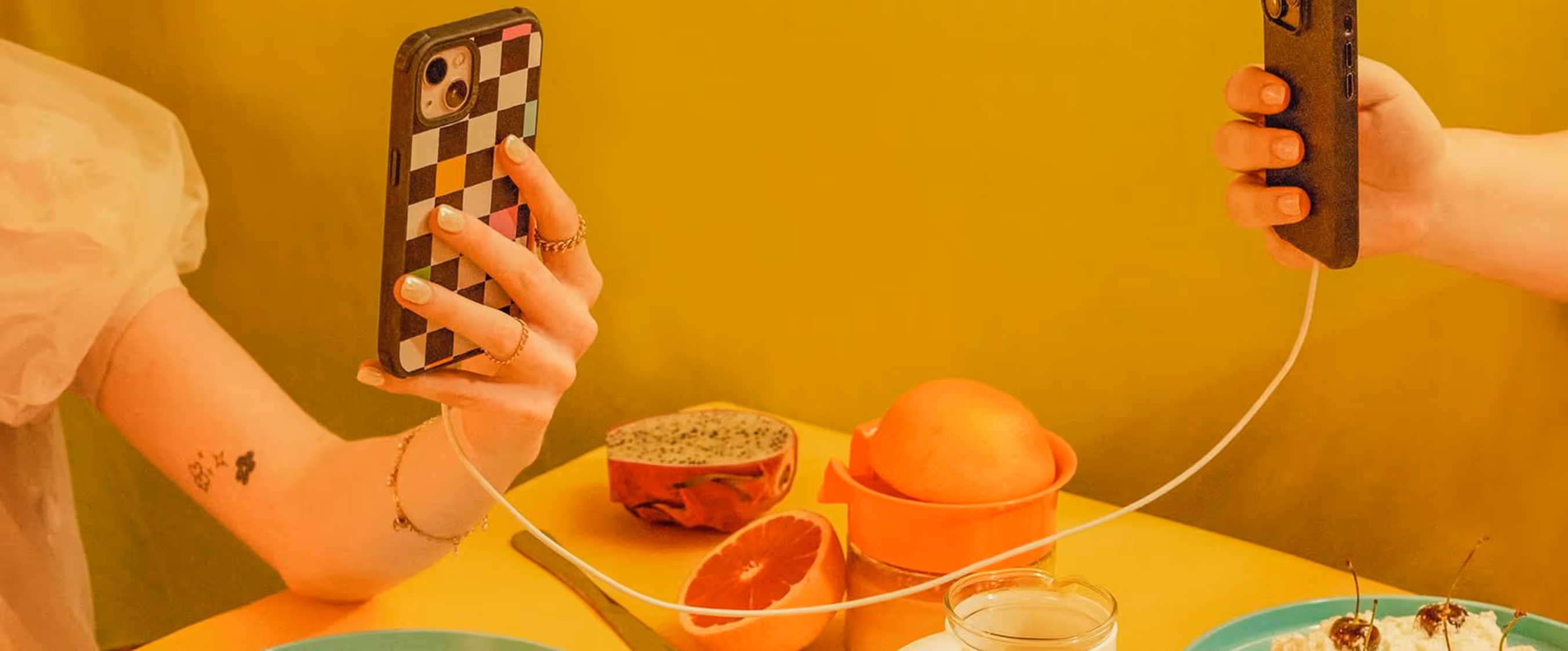 Two hands holding smartphones connected by a white charging cable over a yellow table with fruit and dessert dishes.