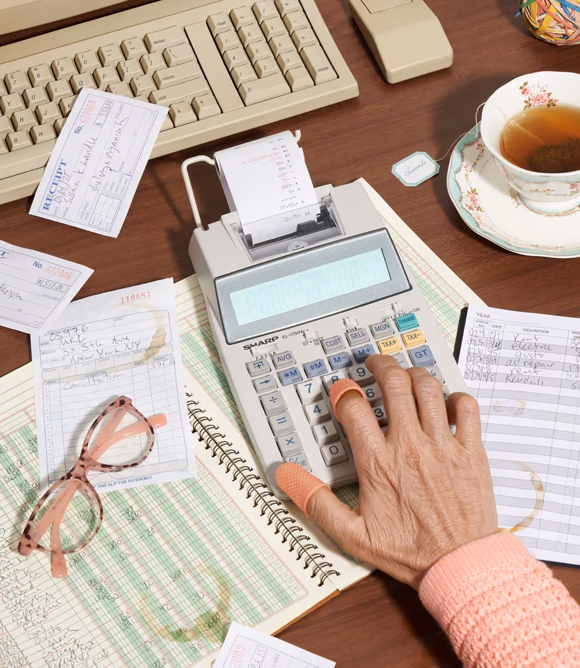 A person’s hand uses a calculator on a desk with a ledger, receipts, glasses, a pen, and a cup of tea beside a computer keyboard.