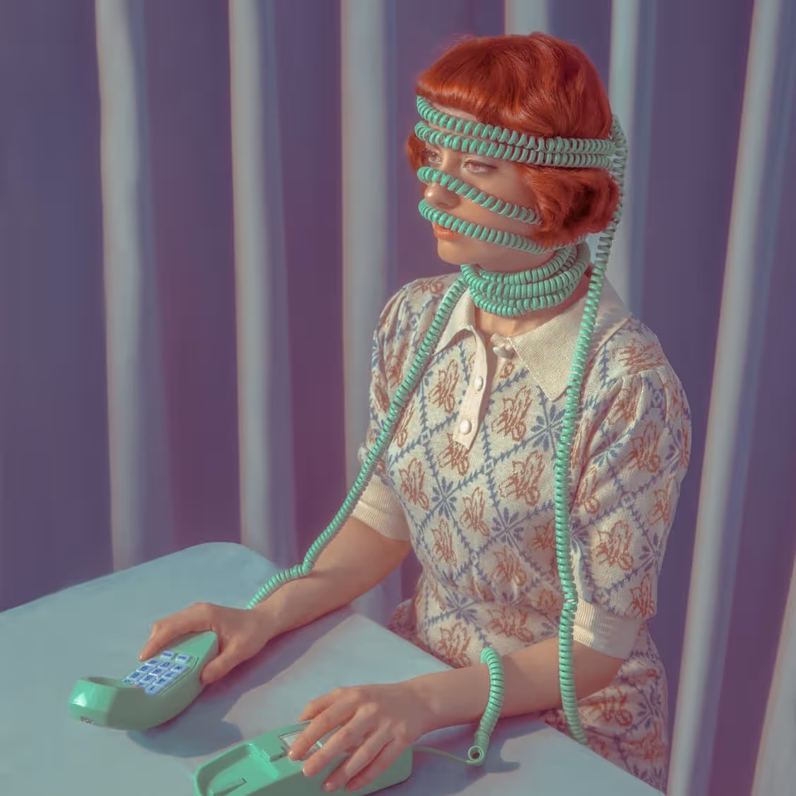 Woman with red hair wrapped in coiled turquoise telephone cords around her head, face, and neck against a purple curtain backdrop.