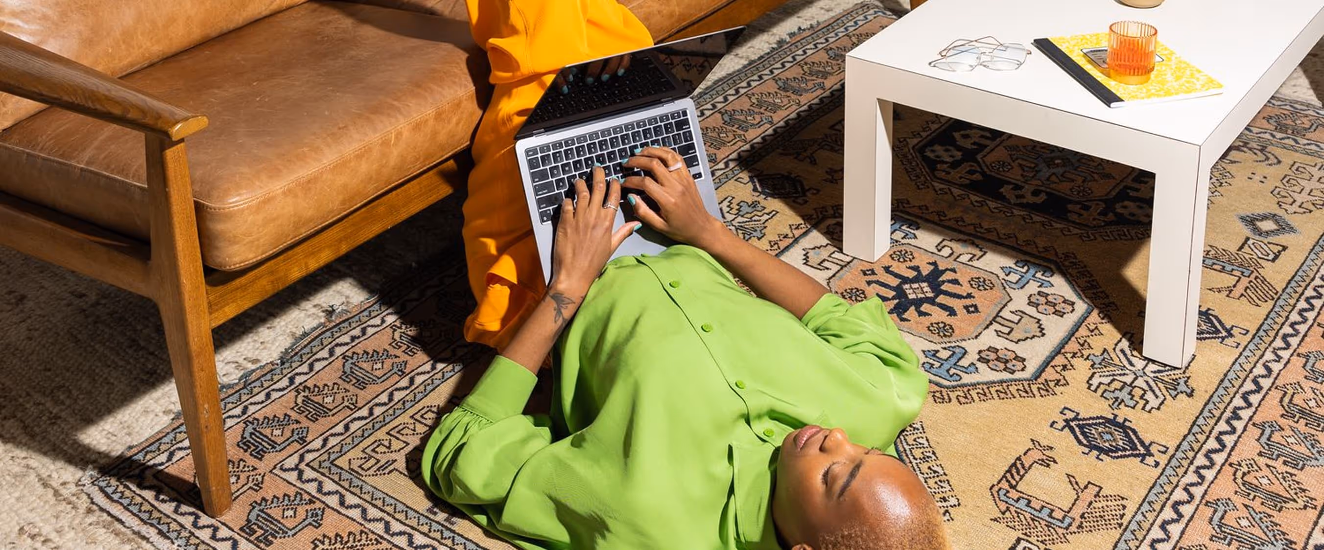 Person in a bright green shirt and orange pants lying on a patterned rug typing on a laptop near a brown leather couch and white coffee table.
