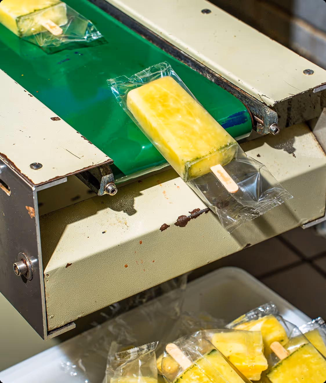 Yellow popsicle in clear plastic wrapper on a green conveyor belt in a manufacturing setting.