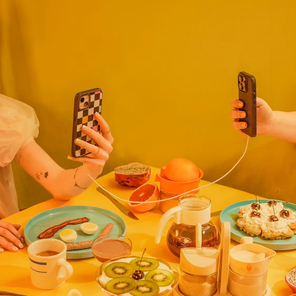 Two hands holding smartphones connected by a white charging cable over a yellow table with fruit and dessert dishes.