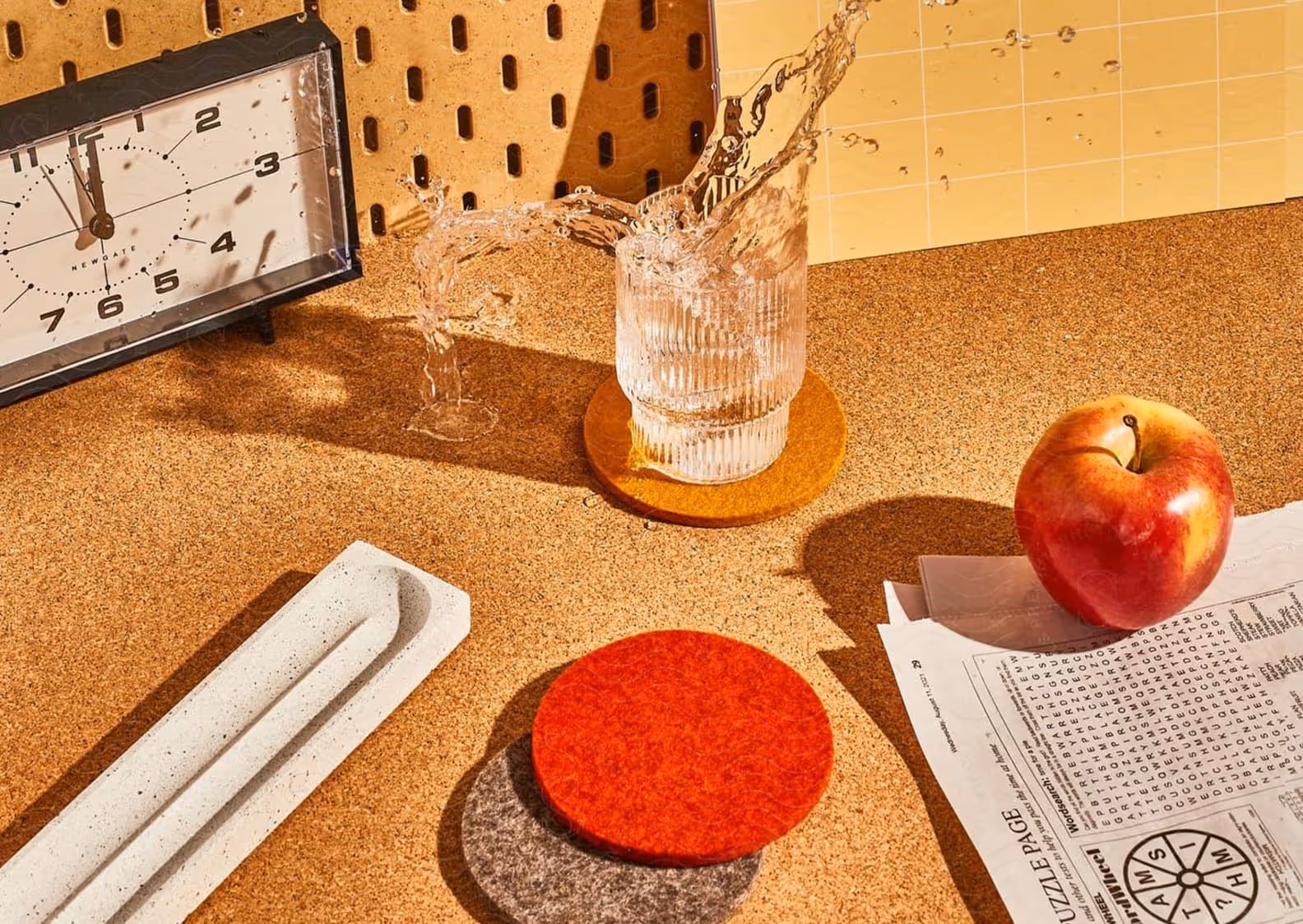 A glass of water spilling over on an orange coaster, next to a red apple on newspaper, a long white tray, and a clock showing 11:58 on a cork surface.