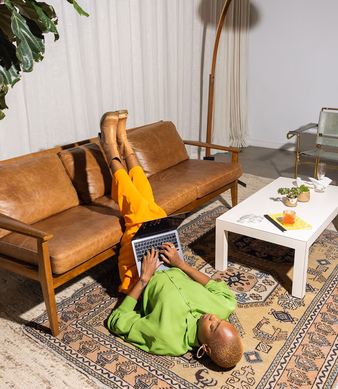 Person in a bright green shirt and orange pants lying on a patterned rug typing on a laptop near a brown leather couch and white coffee table.