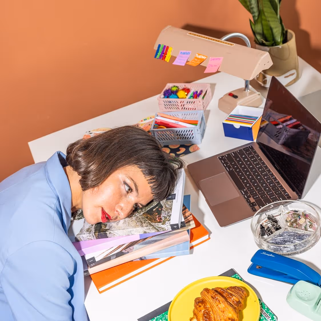 Woman with short hair resting her head on a stack of magazines on a desk beside a laptop and office supplies.