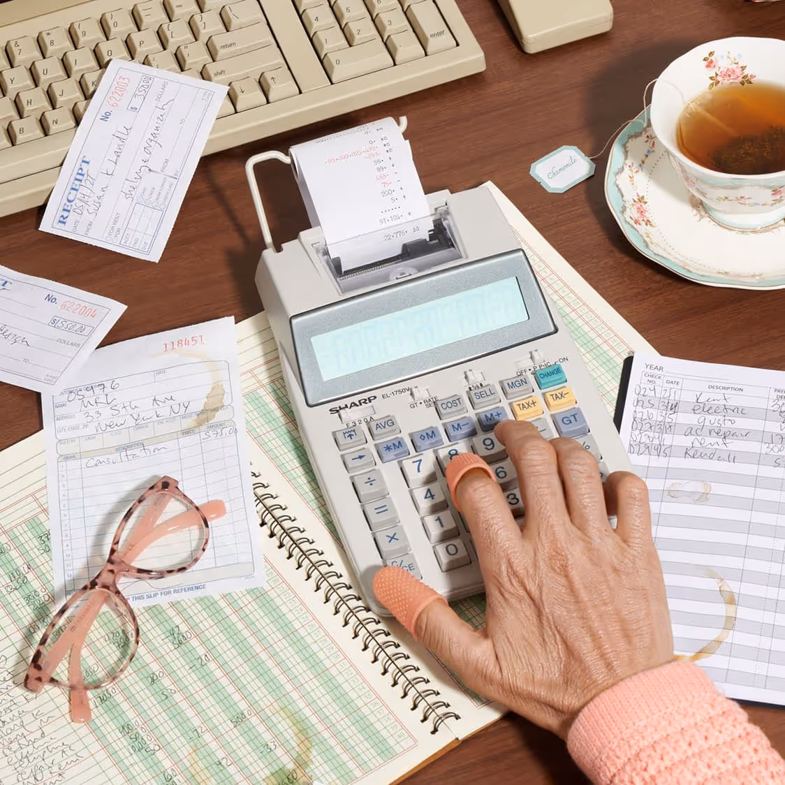 A person’s hand uses a calculator on a desk with a ledger, receipts, glasses, a pen, and a cup of tea beside a computer keyboard.