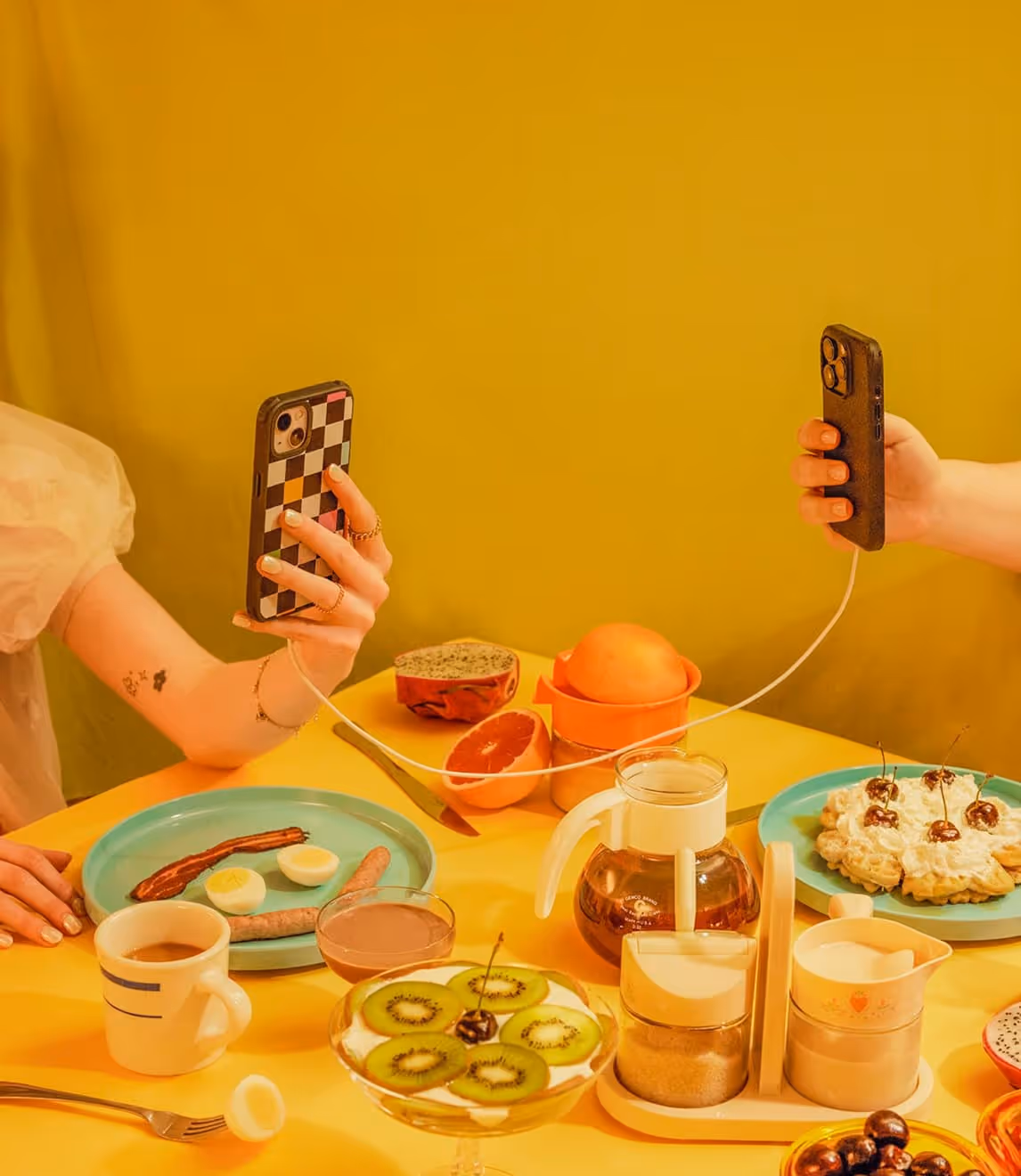 Two hands holding smartphones connected by a white charging cable over a yellow table with fruit and dessert dishes.