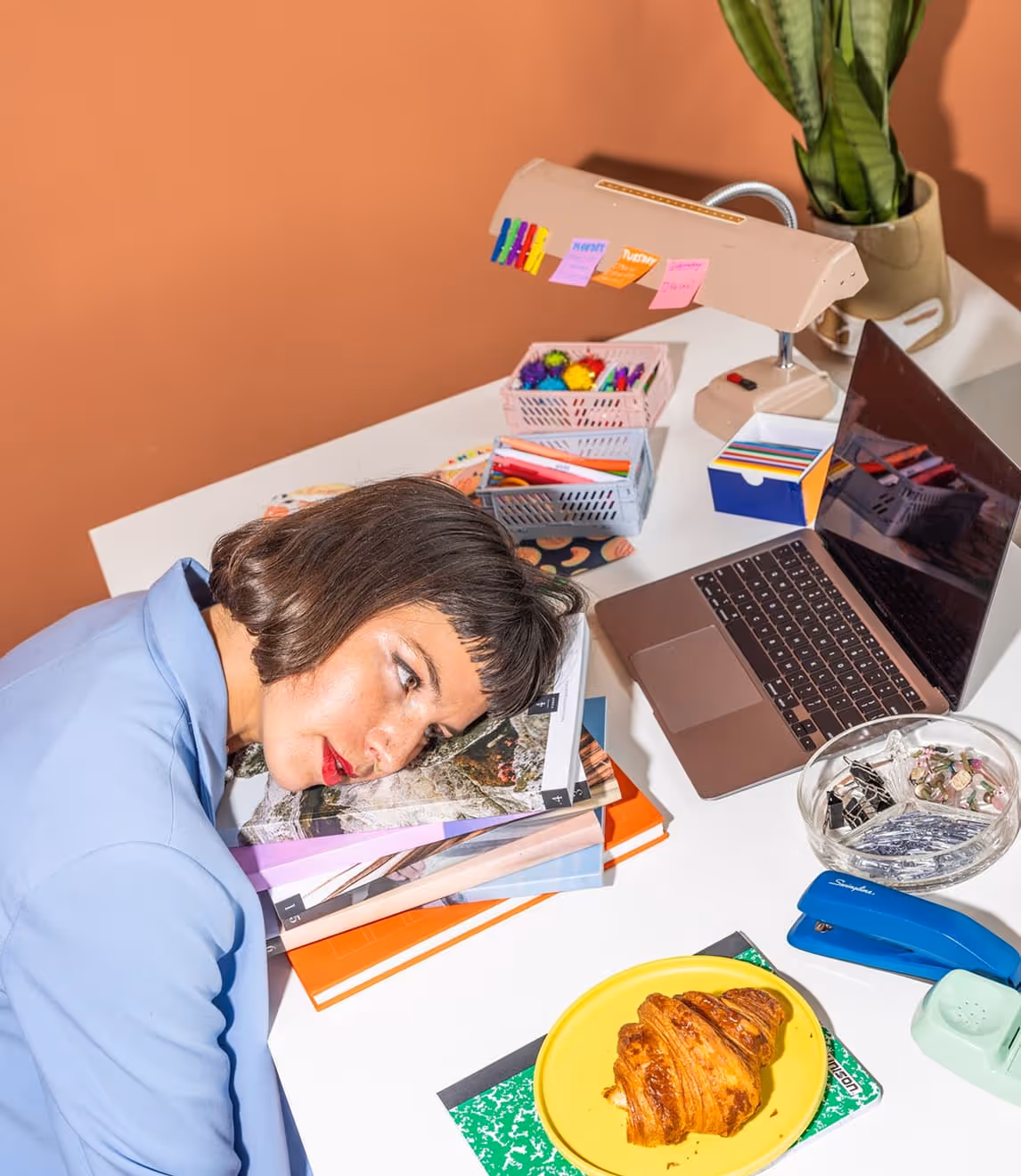 Woman with short hair resting her head on a stack of magazines on a desk beside a laptop and office supplies.