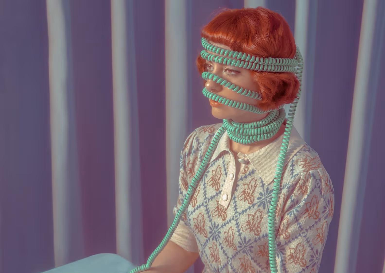 Woman with red hair wrapped in coiled turquoise telephone cords around her head, face, and neck against a purple curtain backdrop.