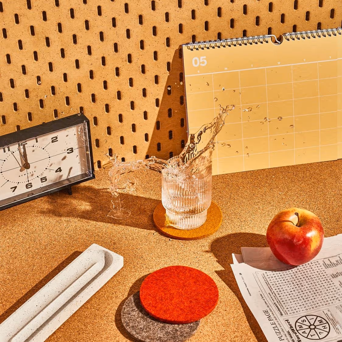 A glass of water spilling over on an orange coaster, next to a red apple on newspaper, a long white tray, and a clock showing 11:58 on a cork surface.