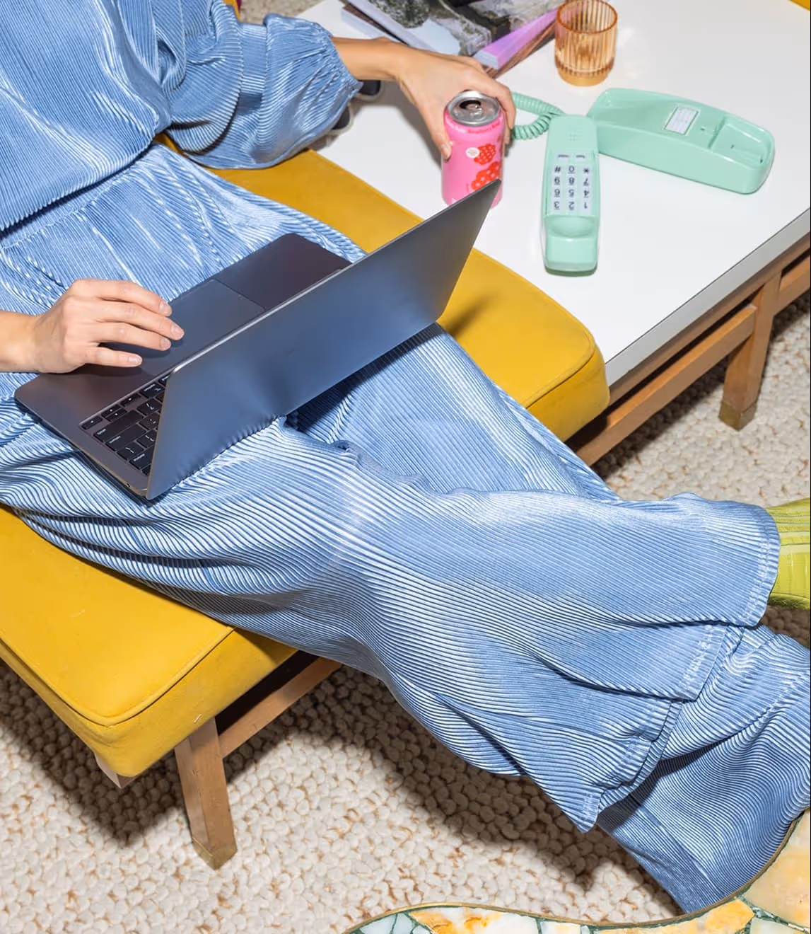 Person in a blue pleated outfit sitting on a yellow bench using a laptop and holding a pink soda can near a white table with a mint green vintage telephone.
