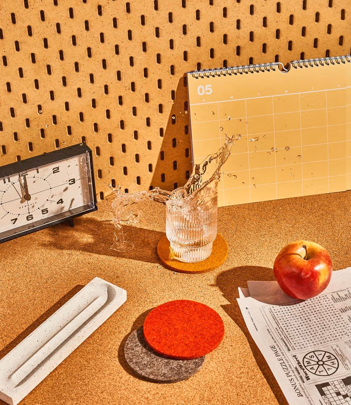 A glass of water spilling over on an orange coaster, next to a red apple on newspaper, a long white tray, and a clock showing 11:58 on a cork surface.